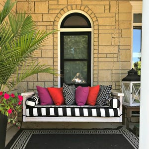 May include: A white porch swing with black and white striped cushions and pillows in red, pink, and black patterns. The swing is hanging from a chain and is in front of a brick wall with a window.
