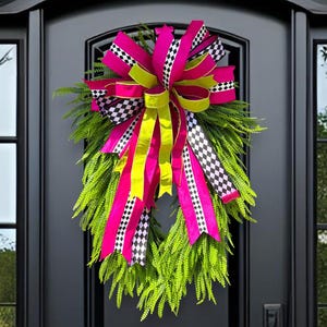 May include: A vibrant green fern wreath adorned with a large bow. The bow features bright pink, yellow, and black and white checkered ribbons. The wreath hangs on a black front door with glass panels.