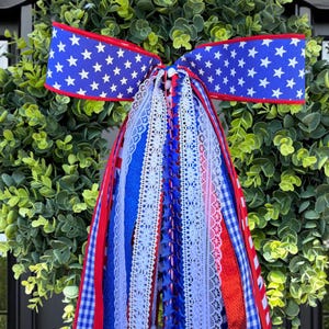 May include: A decorative bow featuring a blue ribbon with white stars and long, cascading streamers in red, white, and blue. The streamers include various textures such as white lace, blue gingham, solid red, and blue ribbons with star patterns.