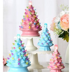 May include: Four ceramic Christmas trees in shades of pink and blue, adorned with colorful lights. The trees vary in size and are displayed on white cake stands, creating a festive holiday scene.
