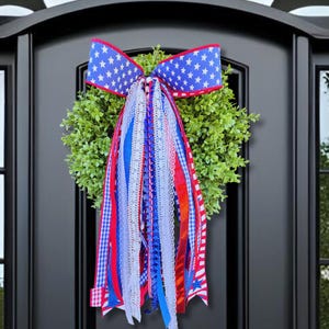 May include: A round green boxwood wreath adorned with a large bow featuring a blue background with white stars. Cascading from the bow are multiple ribbons in red, white, and blue, including lace, gingham, and solid textures, creating a patriotic display.