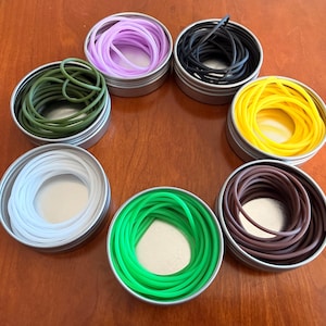 Puede incluir: Un surtido de bandas elásticas de colores en pequeñas latas redondas de metal. Las bandas son de varios colores, incluyendo verde, amarillo, negro, morado, blanco y marrón. Las latas están dispuestas en un patrón circular sobre una superficie de madera.