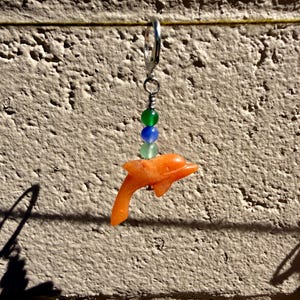 May include: Orange dolphin charm earring with a silver hoop. The earring features a small orange dolphin pendant, with green and blue beads. The earring is hanging against a textured, light brown wall.