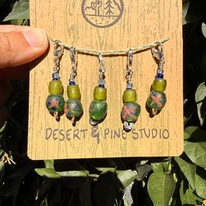 Puede incluir: Un conjunto de cinco pendientes hechos a mano con cuentas verdes y rojas estampadas. Los pendientes están sujetos a una tarjeta marrón con el texto "Desert & Pine Studio" y un logotipo de desierto y pino.