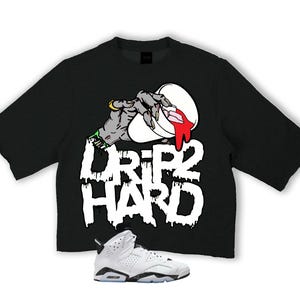 Könnte beinhalten: Schwarzes T-Shirt mit einer Grafik einer Zombiehand, die rote Flüssigkeit aus einem weißen Behälter gießt, mit den Worten "DRIP HARD" in tropfenden weißen Buchstaben. Ein weißer und schwarzer Sneaker befindet sich unten.