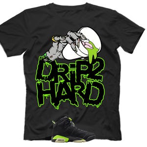 Könnte beinhalten: Schwarzes T-Shirt mit einer Grafik einer Zombiehand, die grüne Flüssigkeit aus einer weißen Tasse gießt. Der Text "DRIP 2 HARD" ist in grünen, tropfenden Buchstaben gehalten. Unten befindet sich ein schwarzer und grüner Sneaker.