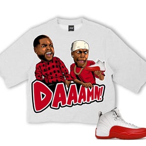 Puede incluir: Una camiseta gris claro con un gr&aacute;fico de dos personajes de dibujos animados y la palabra "DAAAMN!" en rojo. Un par de zapatillas blancas y rojas est&aacute;n en la imagen. La camiseta tiene cuello redondo y mangas cortas.