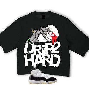 Könnte beinhalten: Schwarzes T-Shirt mit einer Grafik einer Zombiehand, die rote Flüssigkeit aus einem weißen Behälter tropft, mit den Worten "DRIP HARD" in Weiß. Unten ein Paar schwarz-weiße Turnschuhe mit durchscheinender Sohle.