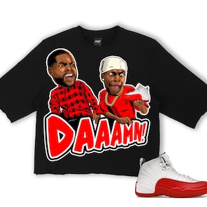 Puede incluir: Camiseta negra con un gr&aacute;fico de dos figuras de dibujos animados y la palabra "DAAAMN!" en rojo. Una zapatilla Air Jordan 12 blanca y roja est&aacute; en la esquina inferior derecha. La camiseta tiene un dobladillo sin rematar.