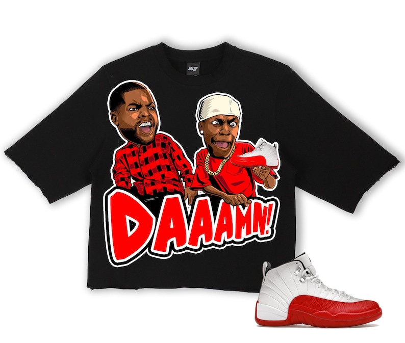 Puede incluir: Camiseta negra con un gr&aacute;fico de dos figuras de dibujos animados y la palabra "DAAAMN!" en rojo. Una zapatilla Air Jordan 12 blanca y roja est&aacute; en la esquina inferior derecha. La camiseta tiene un dobladillo sin rematar.