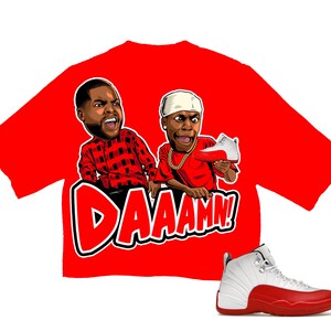 Puede incluir: Camiseta roja con un gr&aacute;fico de dibujos animados de dos hombres y la palabra "DAAAMN!". Una zapatilla Air Jordan 12 blanca y roja tambi&eacute;n est&aacute; en la imagen. La camiseta presenta un dise&ntilde;o audaz y un tema l&uacute;dico.