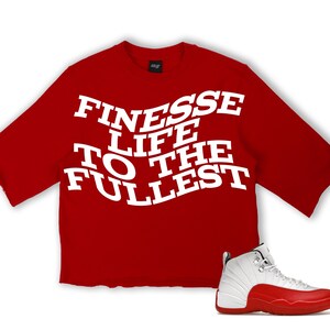 Puede incluir: Una camiseta roja de manga corta con el texto "FINESSE LIFE TO THE FULLEST" en blanco. Una zapatilla deportiva blanca y roja est&aacute; colocada a la derecha de la camiseta. La zapatilla tiene suela roja y parte superior blanca.