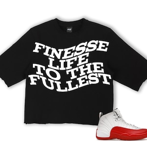 Puede incluir: Camiseta negra de manga corta con el texto blanco "FINESSE LIFE TO THE FULLEST". Una zapatilla deportiva blanca y roja est&aacute; colocada en la parte inferior derecha. La camiseta tiene un dobladillo sin rematar.