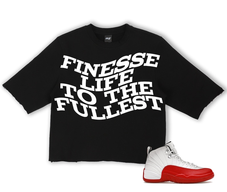 Puede incluir: Camiseta negra de manga corta con el texto blanco "FINESSE LIFE TO THE FULLEST". Una zapatilla deportiva blanca y roja est&aacute; colocada en la parte inferior derecha. La camiseta tiene un dobladillo sin rematar.