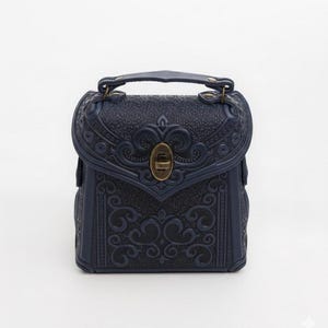 May include: A navy blue, small, square handbag with a handle and ornate embossed detailing. The bag features a decorative clasp and metal hardware. The design includes floral and scroll patterns. The bag is approximately 18 cm tall.