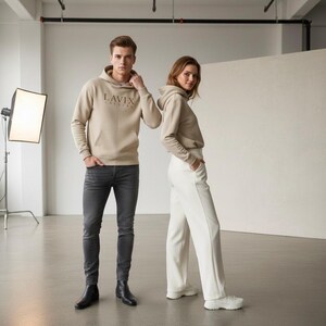 May include: Two people wearing matching beige hoodies with the text "LAVIX CULTURE" printed on the front. One person is wearing dark gray jeans and black boots, while the other is wearing white pants and white sneakers. The background is a studio setting with a large white backdrop.