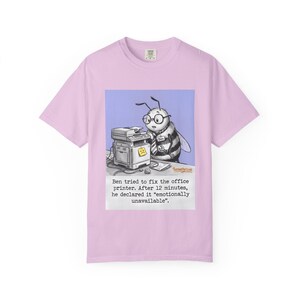 May include: A light pink t-shirt featuring a cartoon of a bee with glasses, working on a printer. The text reads, "Ben tried to fix the office printer. After 12 minutes, he declared it 'emotionally unavailable.'"