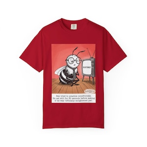 May include: Red t-shirt featuring a cartoon bee wearing glasses and a tie, sitting in front of a television with the word "ENLIGHTENMENT" on the screen. The text below reads, "Ben tried to practice mindfulness..."