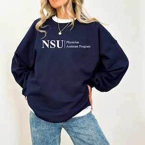 Könnte beinhalten: Marineblaues Sweatshirt mit dem weißen Text "NSU Physician Assistant Program". Das Sweatshirt hat lange Ärmel und einen Rundhalsausschnitt. Die Person trägt ein weißes Unterhemd und blaue Jeans.