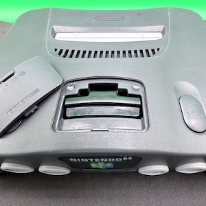 May include: A gray Nintendo 64 game console with a matching gray expansion pack. The console features a slot for game cartridges, a reset button, and the Nintendo 64 logo. The expansion pack is labeled "MEMORY EXPANSION".