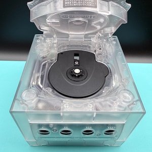 May include: A clear, transparent video game console with an open lid, revealing a black disc tray. The console has multiple ports and the text "テイスクやメモリーカードは最後まで押し込んでください。! レンズに触れないでください。" is visible on the lid.