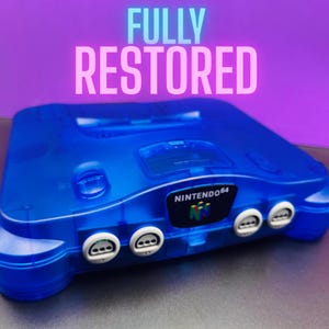 May include: A transparent blue Nintendo 64 console with the text "FULLY RESTORED" in pink and blue. The console has the Nintendo 64 logo and four controller ports.
