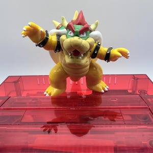May include: A yellow Bowser figurine with a red and green shell, red horns, and black wristbands. The figurine is posed with arms outstretched, standing on a red, transparent plastic surface. The figure is a character from the Super Mario video game series.