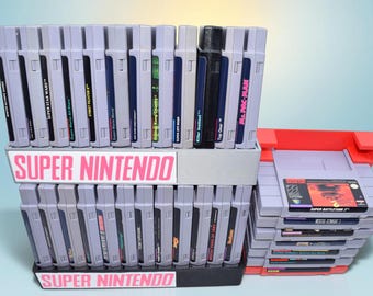 May include: A collection of Super Nintendo video game cartridges organized in two gray plastic display stands with pink Super Nintendo labels. A separate stack of cartridges sits to the right in a red plastic holder, featuring titles like Super Battletank 2.