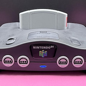 May include: A gray Nintendo 64 video game console with a game cartridge inserted. The console has a curved design with four controller ports and the Nintendo 64 logo on the front. A blue power light is illuminated.