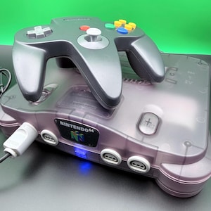 May include: A translucent purple Nintendo 64 console with a matching controller. The controller has a gray directional pad, a white analog stick, and colored buttons. The console has the Nintendo 64 logo and a power button.