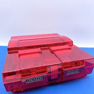 May include: A transparent red video game console with visible internal components. The console has a rectangular shape with multiple ports and a top loading slot. The design includes a series of rectangular and square shapes. The console is set against a blue surface.