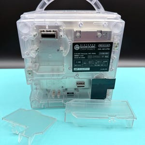 May include: A clear, transparent Nintendo GameCube console with a handle, showing internal components. The console has a rectangular shape with a removable panel and the Nintendo GameCube logo. The model number is DOL-001(JPN).