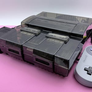May include: A translucent black Super Nintendo Entertainment System console featuring a top-loading cartridge slot, two front controller ports, a power adapter, a grey controller with purple buttons, and a set of yellow, white, and red AV cables.