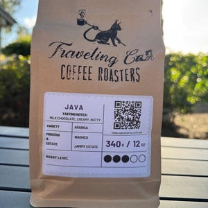 May include: A brown paper bag of coffee from Traveling Cat Coffee Roasters. The bag features a black cat logo and the text "JAVA". Tasting notes include milk chocolate, creamy, and nutty. The bag contains 12 oz of coffee.