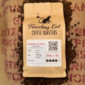 May include: A brown paper bag of coffee beans labeled "Traveling Cat Coffee Roasters." The bag, surrounded by coffee beans, features the text "Wander Blend" and details including tasting notes, variety, origin, and roast level. The bag contains 12 oz of coffee.