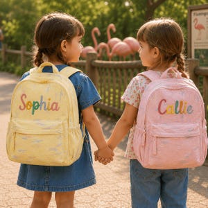 Könnte beinhalten: Zwei Rucksäcke mit einer strukturierten, mehrfarbigen Pinselstrich-Optik. Der gelbe Rucksack trägt den Namen Sophia in Regenbogenfarben gestickt, während der rosa Rucksack den Namen Callie in der gleichen regenbogenfarbenen Stickerei zeigt.