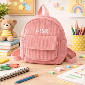 Może przedstawiać: Różowy sztruksowy plecak z białym haftem z imieniem "Lisa". Mały plecak ma przednią kieszeń i górny uchwyt. Tło obejmuje pluszowego misia, ołówki i notatnik.