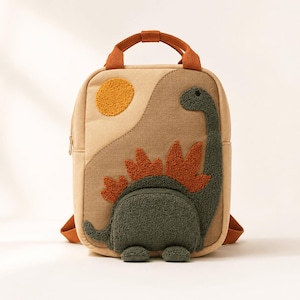 May include: A beige canvas backpack featuring a textured, dark green dinosaur appliqué with orange spikes on the front pocket. A yellow, fuzzy circular sun is positioned in the upper left corner. The bag includes two top handles and adjustable straps.