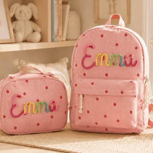 May include: A pink corduroy backpack and matching lunch bag, both featuring a red polka dot pattern and the name Emma embroidered in colorful, textured yarn. Each bag has a top handle, a front zippered pocket, and white tassel zipper pulls.