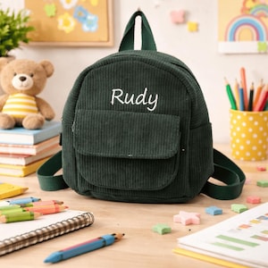 Op de afbeelding: Een donkergroene corduroy rugzak met de naam "Rudy" in het wit geborduurd. De kleine rugzak heeft een voorvak en een handvat aan de bovenkant. De tas staat op een houten oppervlak met schoolspullen en een teddybeer op de achtergrond.