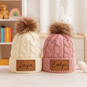 May include: Two cable-knit bobble hats, one cream and one pink, each with a brown faux fur pompom. The cream hat has a brown leatherette patch with the name "Layia". The pink hat has a patch with the name "Evelyn".