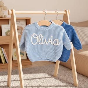Op de afbeelding: Lichtblauwe gebreide trui met de naam "Olivia" in witte letters. Een effen blauwe trui hangt ernaast. Beide hangen aan houten kleerhangers. Kinderkleding collectie.