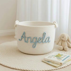 Op de afbeelding: Een witte touwmand met de naam "Angela" in lichtblauw schrift. De mand heeft twee touwhandvatten en staat op een rond, crèmekleurig tapijt. Een knuffelkonijn en een kinderboek liggen in de buurt.