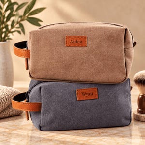 Könnte beinhalten: Zwei rechteckige Kulturbeutel mit Seitengriffen, einer aus beigem Canvas und einer aus dunkelgrauem Canvas. Jede Tasche hat einen rechteckigen braunen Lederaufnäher auf der Vorderseite. Die beige Tasche ist mit dem Namen Aiden und die graue mit Wyatt geprägt.