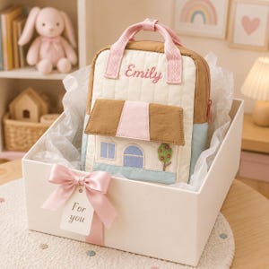 May include: A quilted backpack featuring a house design with a brown roof, light blue walls, and a pink handle. The name Emily is embroidered in pink script on the top. The backpack is presented in a white gift box with a pink ribbon and a tag reading For you.