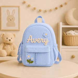 Può includere: Uno zaino in velluto a coste azzurro con il nome Avery ricamato in corsivo color crema sulla tasca anteriore. La borsa è decorata con piccole applicazioni floreali blu all'uncinetto con steli verdi e una maniglia superiore per il trasporto.