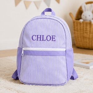 Può includere: Uno zaino a righe verticali color lavanda e bianco con una tasca frontale con cerniera e il nome CHLOE ricamato in filo viola al centro. Lo zaino include una maniglia superiore, spallacci regolabili e pannelli laterali in tinta unita.