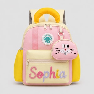 Puede incluir: Una mochila de color rosa pastel y amarillo con un asa superior amarilla brillante, un bolsillo frontal con el nombre Sophia bordado en letra cursiva colorida y un estuche circular rosa desmontable con una cara de conejo y bigotes.
