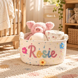 Peut inclure: Un grand panier de rangement rond en corde blanc cassé portant le prénom Rosie en lettres de laine texturées et colorées. Le panier est orné de petits points de laine multicolores et de fleurs en laine en relief roses, bleues et jaunes.