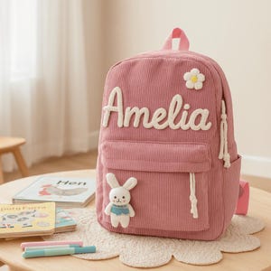 Peut inclure: Sac à dos en velours côtelé rose avec le nom "Amelia" en écriture blanche. Comprend une marguerite blanche et une applique de lapin. Comprend une poche avant, une poche latérale et des détails de cordon blancs. Le sac à dos est posé sur une table en bois.